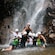 Excited men and women, wearing helmets, pose for a picture under a waterfall